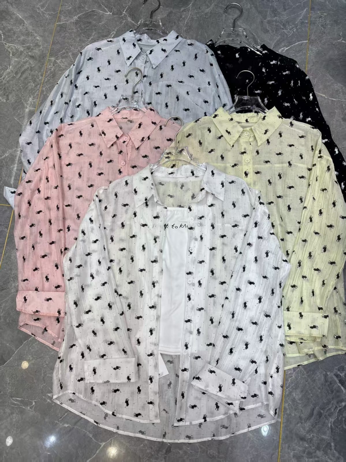 Women's polo pony print sheer shirt in 5 colours — white, light blue, blush pink, butter yellow and black — displayed on hangers on marble floor