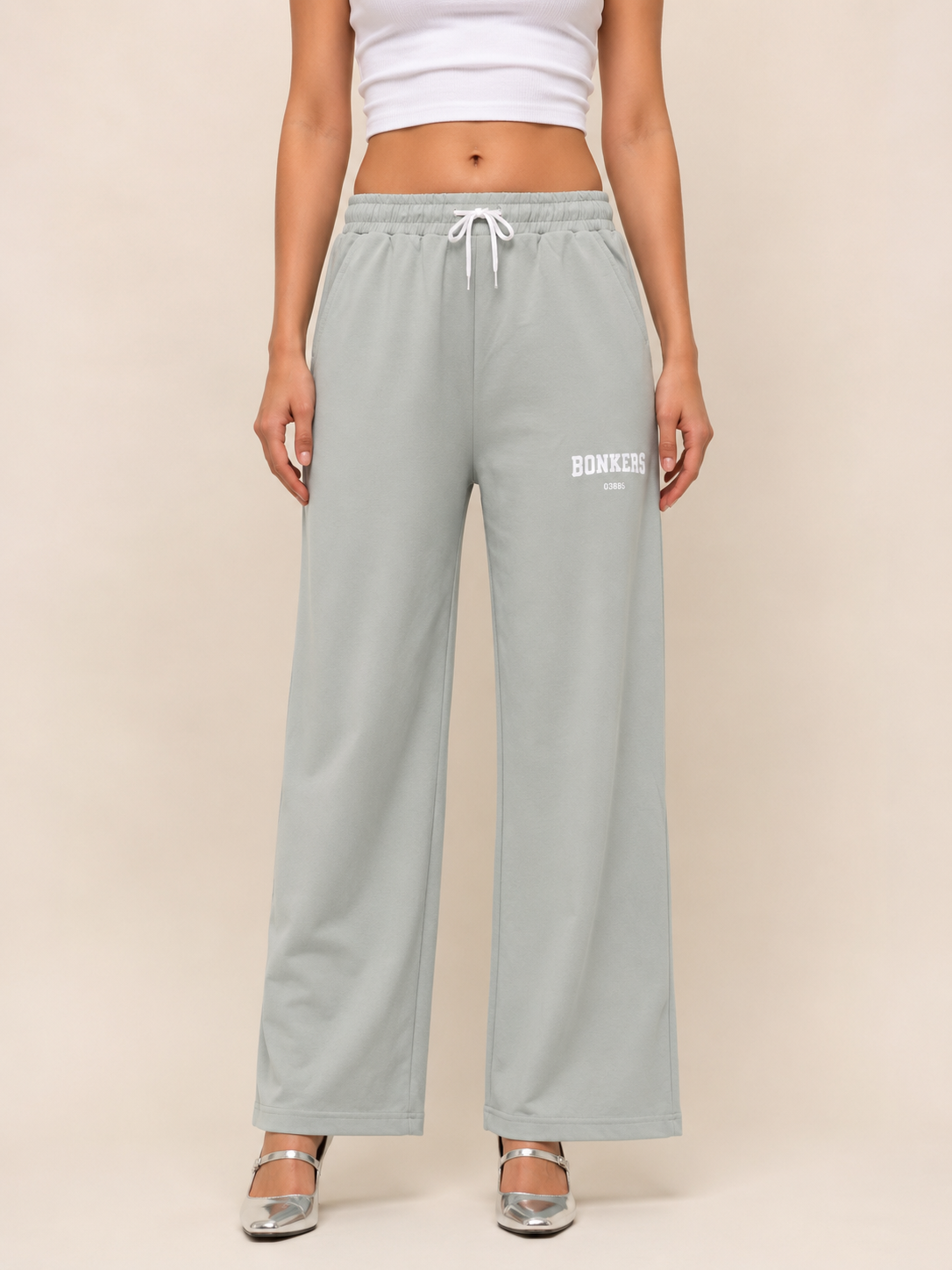 A model is wearing light sage green wide-leg sweatpants with a white drawstring and 'BONNIE' printed in white on the left thigh, paired with a white crop top and silver heels, set against a solid beige background.