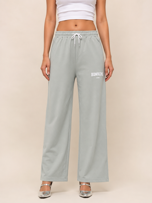 A model is wearing light sage green wide-leg sweatpants with a white drawstring and 'BONNIE' printed in white on the left thigh, paired with a white crop top and silver heels, set against a solid beige background.